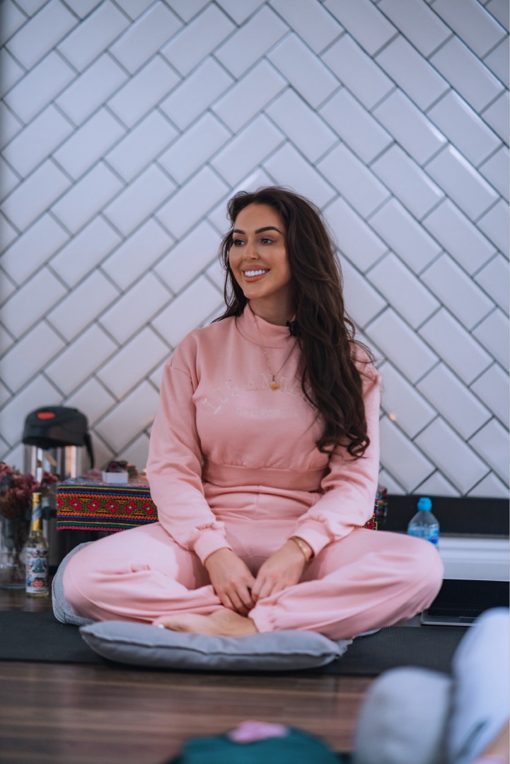Shona Gray seated cross-legged, smiling warmly in a pink outfit against a herringbone wall